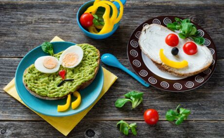 Farfurie albastră așezată pe un șervețel galben, cu tartină cu avocado și 2 jumătăți de ou, decorată cu ardei galben și busuioc, castron cu legume și o farfurie negru cu alb și o tartină cu brânză, decorată cu ardei și roșii, așezate pe o masă din lemn, decorată cu câteva frunze de busuioc, o roșie și o linguriță albastră, ilustrând top 7 alimente sănătoase pentru copii