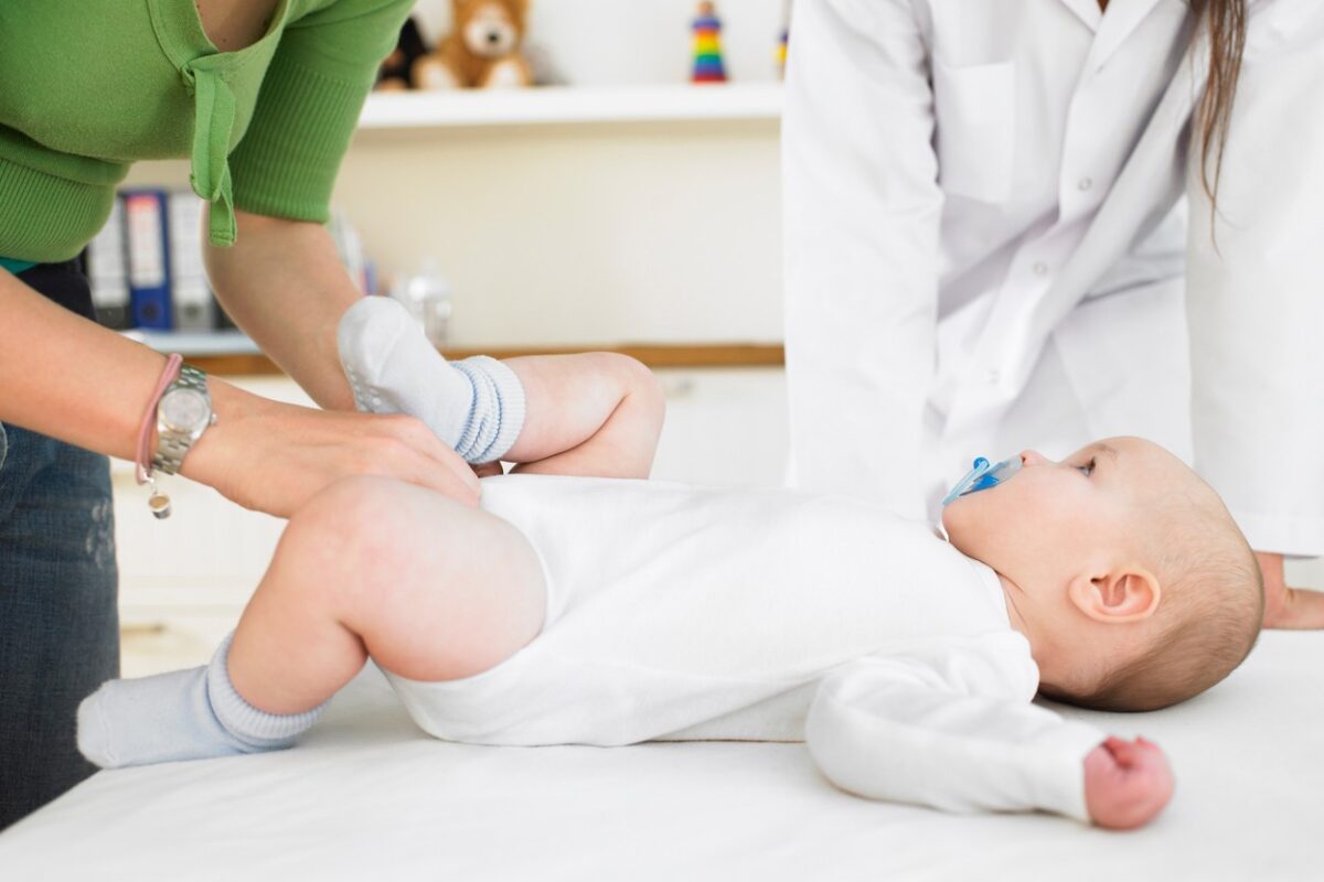 Un bebeluș care stă pe masa din cabinetul medicului și este dezbrăcat de mamă pentru un control pediatric