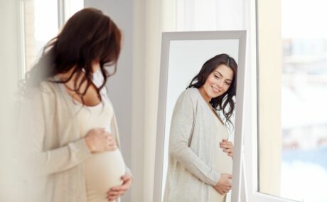 Femeie însărcinată, îmbrîcată cu o rochie bej, cu un cardigan bej pe deasupra, care se uită în oglindă, în dormitor, lângă fereastră și zâmbește, ilustrând și îți ține burta cu mâinile, ilustrând o „voce interioară“ pozitivă în perioada sarcinii