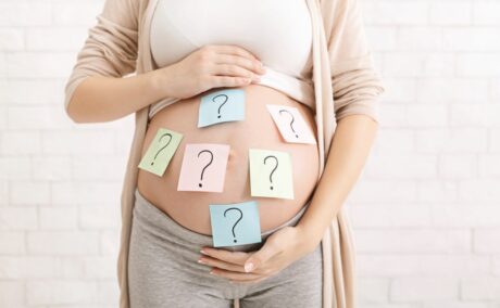 Femeie însărcinată îmbrăcată cu un maiou și cardigan bej și colanți gri, are burta descoperită și postituri colorate cu semnul întrebării pe fiecare dintre ele lipite, care ilustrează alegerea numeleui bebelușului