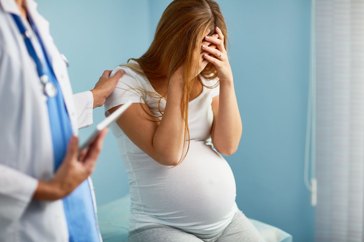 Femeie însărcinată, îmbrăcată cu tricou alb și colanți gri, care stă așezată în fund pe pat, cu mâinile acoperindu-și fața, iar medicul ține un carnețel în mână și îi ține o mână pe umăr, ilustrând anencefalia