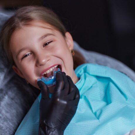 Fetiță la stomatolog, pe scaun, cu un șervet albastru, din hârtie, la gât, iar medicu încearcă să o ajute să rezolve problema cu bruxismul, folosind o gutieră