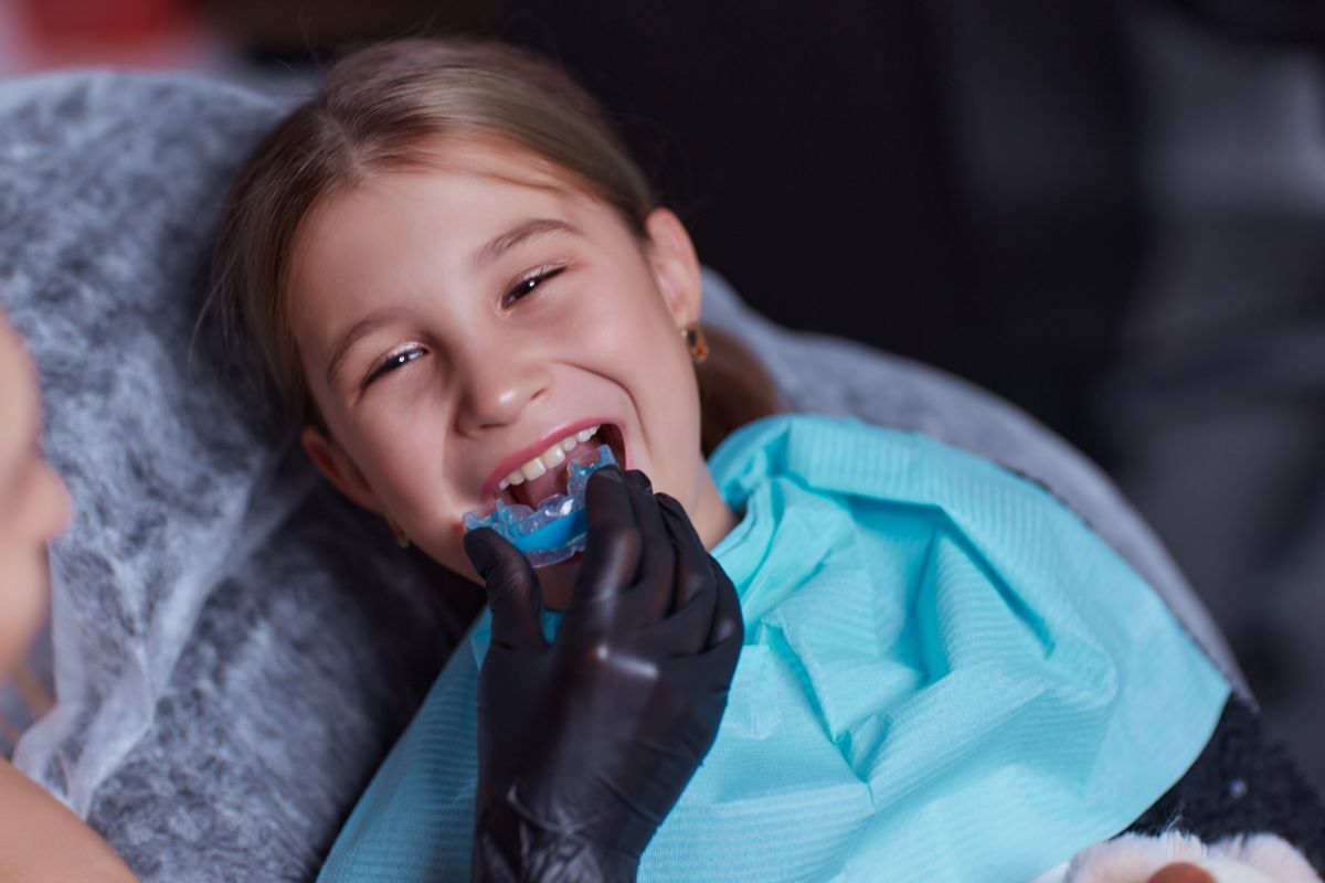 Fetiță la stomatolog, pe scaun, cu un șervet albastru, din hârtie, la gât, iar medicu încearcă să o ajute să rezolve problema cu bruxismul, folosind o gutieră