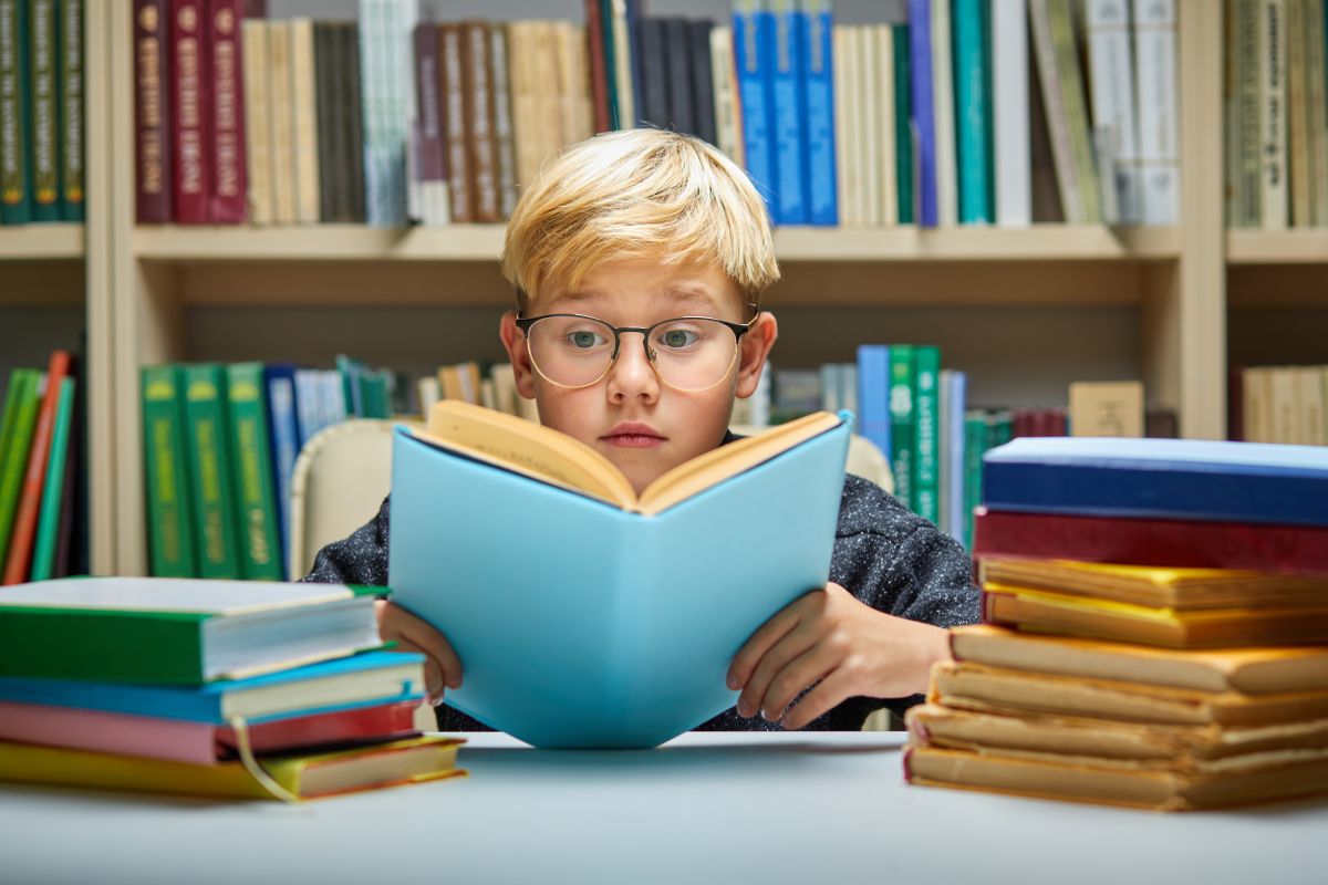 Copil care stă asezat la masă, într-o bibliotecă cu o carte cu coperți albastre și multe cărți în stânga și dreapta mesei, ilustrând 5 semne că ai un copil supradotat