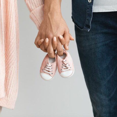 Cuplu de tineri, ea îmbrăcată cu un pulover roz-somon, tricou alb și jeanși bej, iar el cu cu tricou alb și cămașă și pantaloni de jeans, care se țin de mână și țin o pereche de pantofi de bebeluș, roz, ilustrând cum să rămâi cât mai repede însărcinată