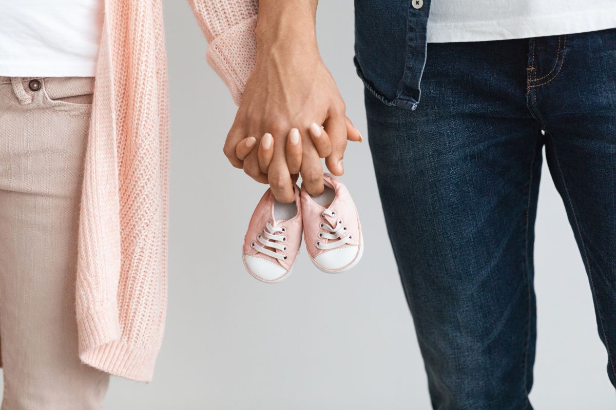 Cuplu de tineri, ea îmbrăcată cu un pulover roz-somon, tricou alb și jeanși bej, iar el cu cu tricou alb și cămașă și pantaloni de jeans, care se țin de mână și țin o pereche de pantofi de bebeluș, roz, ilustrând cum să rămâi cât mai repede însărcinată