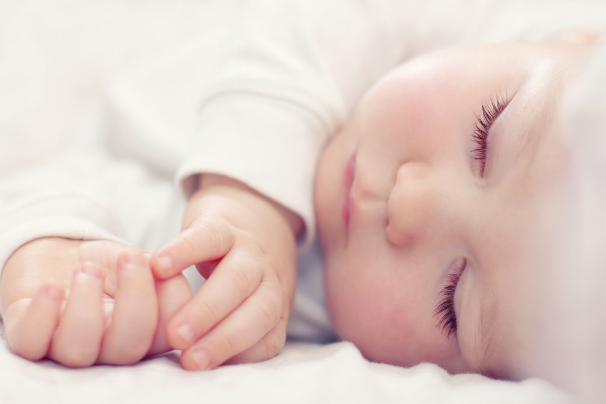 Poză cu bebeluș îmbrăcat cu body alb, care doarme pe-o parte, cu mâinile lângă cap, ilustrând de ce dorm nou-născuții toată ziua