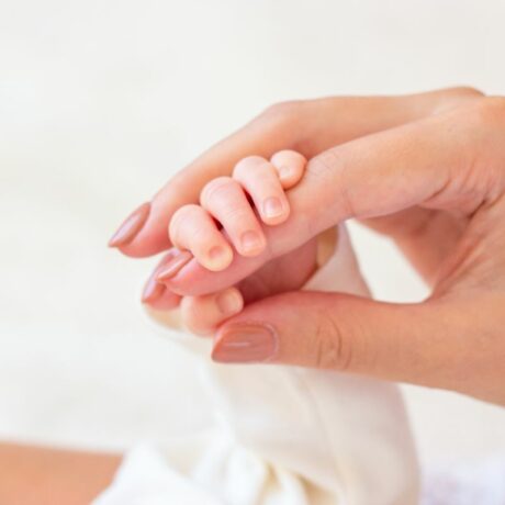 Mâna unei mane, cu unghiile date cu lac maro, care are mânuța unui bebeluș strânsă în jurul degetului arătător, ilutrând de ce îți țin bebelușii pumnii strânși
