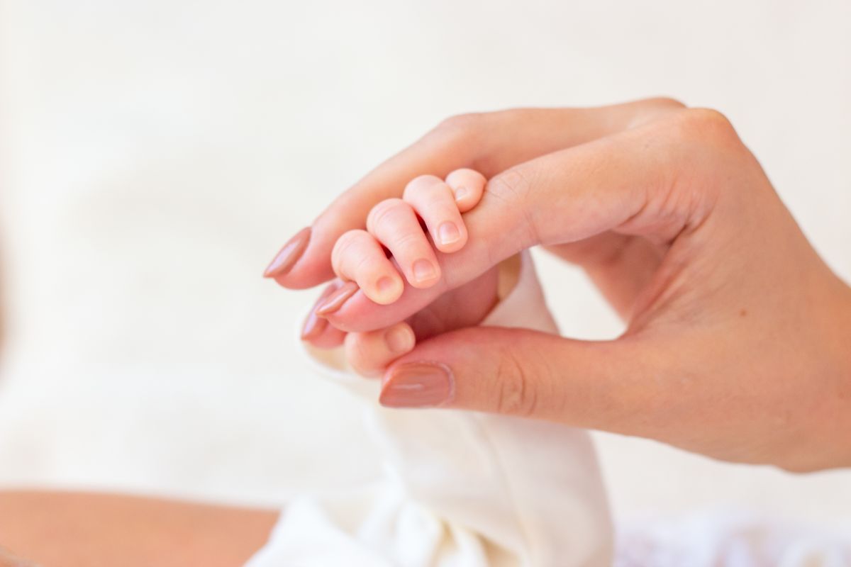 Mâna unei mane, cu unghiile date cu lac maro, care are mânuța unui bebeluș strânsă în jurul degetului arătător, ilutrând de ce îți țin bebelușii pumnii strânși