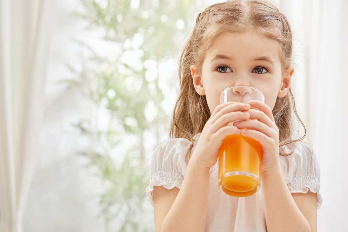 Fetiță, îmbrăcată cu o rochiță albă, care ține în mâini și își duce la gură un pahar cu suc de portocale, ilustrând sucul de fructe