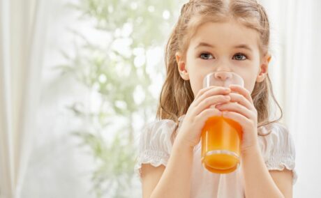 Fetiță, îmbrăcată cu o rochiță albă, care ține în mâini și își duce la gură un pahar cu suc de portocale, ilustrând sucul de fructe