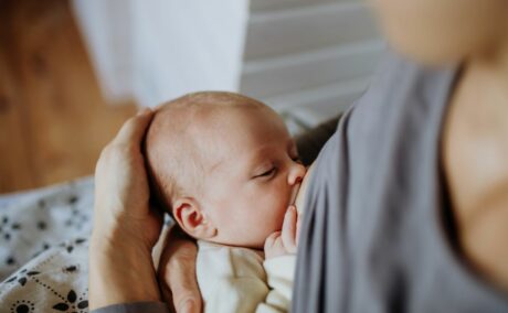 Mămică, care stă așezată, cu bluza gri ridicată în timp ce alăptează bebelușul, ținându-i capul cu mâna și este acoperită pe picioare cu o pătură gri cu motive florale negre, ilustrând dezvoltarea sistemului imunitar al bebelușilor