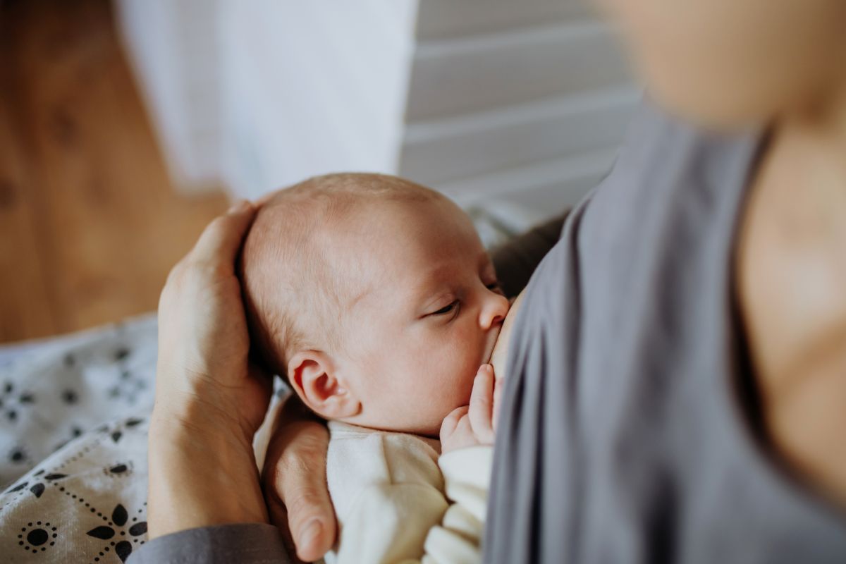 Mămică, care stă așezată, cu bluza gri ridicată în timp ce alăptează bebelușul, ținându-i capul cu mâna și este acoperită pe picioare cu o pătură gri cu motive florale negre, ilustrând dezvoltarea sistemului imunitar al bebelușilor