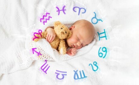 Bebeluș, înfășat într-o păturică albă, cu găuri, stă pe-o parte, ținând în brațe un ursuleț maro, din pluș, stând pe pat, pe o pătură albă, pufoasă, iar în jurul lui apar simbolurile zodiilor cu mov și albastru, ilustrând horoscopul bebelușilor.