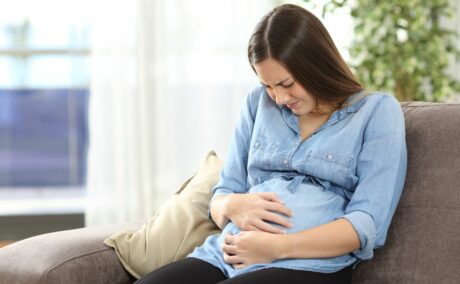 Femeie însărcinată, îmbrăcată cu o bluză din jeans albastru și colanți negri, care se ține cu mîinile de burtă și stă pe o canapea maro cu o pernă bej la spate, ilustrând Infecții care pot provoca malformații congenitale
