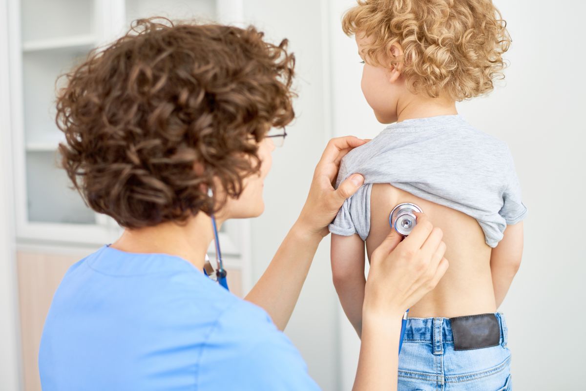 Medic îmbrăcat în halat albastru, cu stetoscopul în urechi, care ascultă plămânii, unui copil, care are tricou gri și jeanși, și stă cu spatele, pentru a verifica pneumonia la copii