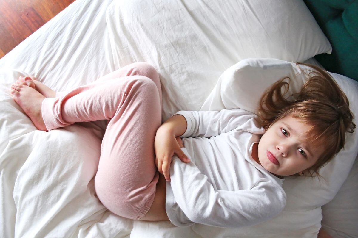 Fetiță cu rotavirus, îmbrăcată cu o bluză de corp albă și colanți roz, care stă în pat, acoperit cu un cearșaf alb, cu genunchii ridicați și se ține de burtă