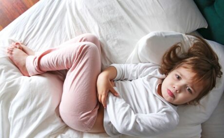 Fetiță cu rotavirus, îmbrăcată cu o bluză de corp albă și colanți roz, care stă în pat, acoperit cu un cearșaf alb, cu genunchii ridicați și se ține de burtă