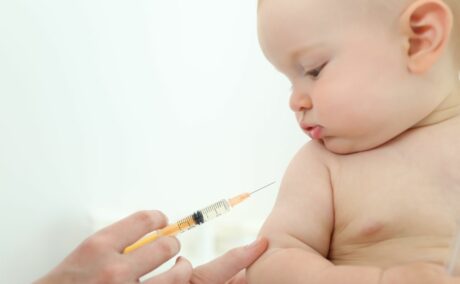 Bebeluș care stă dezbrăcat și se uită la o seringă galbenă, care i se apropie de braț, pentru a primi unul dintre vaccinurile din schema națională de vaccinare a copiilor
