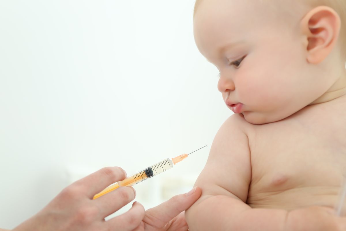 Bebeluș care stă dezbrăcat și se uită la o seringă galbenă, care i se apropie de braț, pentru a primi unul dintre vaccinurile din schema națională de vaccinare a copiilor