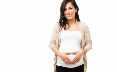 Femeie însărcinată, îmbrăcată cu un top alb și pe deasupra cu un cardigan bej și pantaloni negri, își ține mâinile pe burtă în formă de inimă, ilustrând 12 semne că primul  trimestru de sarcină merge bine