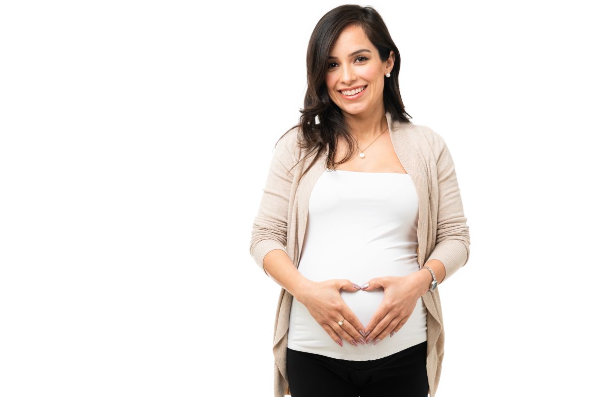 Femeie însărcinată, îmbrăcată cu un top alb și pe deasupra cu un cardigan bej și pantaloni negri, își ține mâinile pe burtă în formă de inimă, ilustrând 12 semne că primul  trimestru de sarcină merge bine