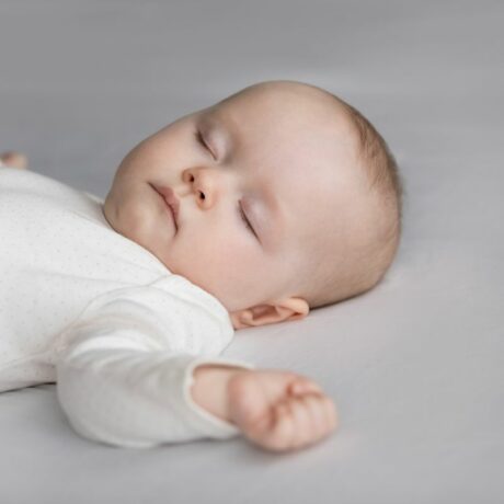 Bebeluș îmbrăcat în body alb, care doarme pe spate, pe un fundal gri