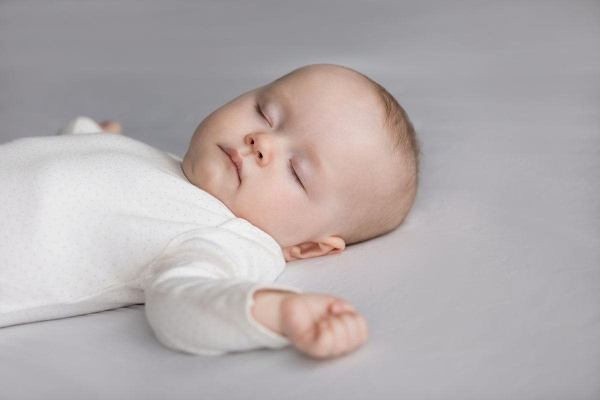 Bebeluș îmbrăcat în body alb, care doarme pe spate, pe un fundal gri