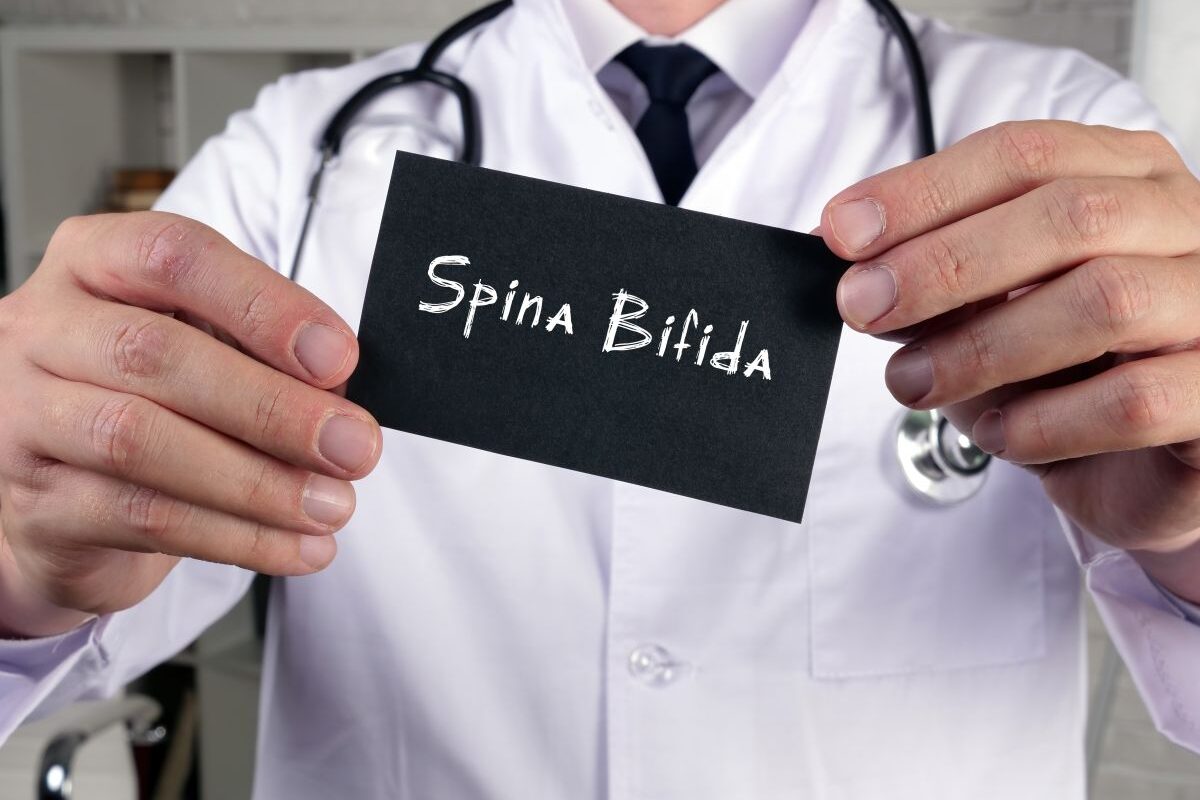 Medic, în halat alb, care are un stetoscop de gât și ține în mână o plăcuță pe care scrie spina bifida