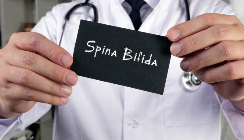 Medic, în halat alb, care are un stetoscop de gât și ține în mână o plăcuță pe care scrie spina bifida