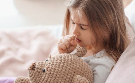 Fetiță care stă în pat, îmbrăcată cu o bluză de pijama gri și ține lângă ea un urs matro, tricotat, și duce pumnul strâns la gură pentru a ilustra tipuri de tuse