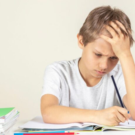 Băiat care stî la un birou și are în față un caiet, stă cu o mână la frunte, ținând în cealaltă mână un creion negru și în fața caietului mai sunt un creion roșu și unul albastru, încercând să-și facă tema, ilustrând tulburările de învățare la copii