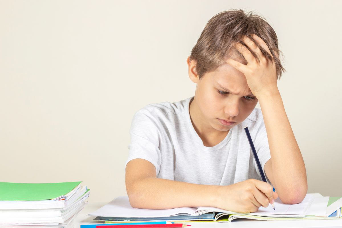 Băiat care stî la un birou și are în față un caiet, stă cu o mână la frunte, ținând în cealaltă mână un creion negru și în fața caietului mai sunt un creion roșu și unul albastru, încercând să-și facă tema, ilustrând tulburările de învățare la copii