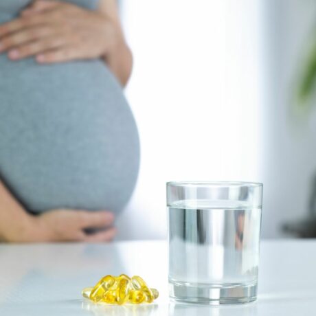 Femeie însărcinată, îmbrăcată într-o rochie mulată, gri, care își ține burta cu mâinile, având în față, pe masă, un pahar cu apă și câteva pastile galbene de vitamina D în sarcină