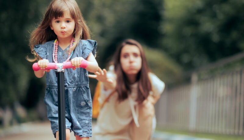 Fetiță, îmbrăcată cu rochiță din jeans, pe trotinetă, în parc, iar mama ei stă în spatele ei și pare foarte îngrijorată, ilustrând 5 temeri ale părinților