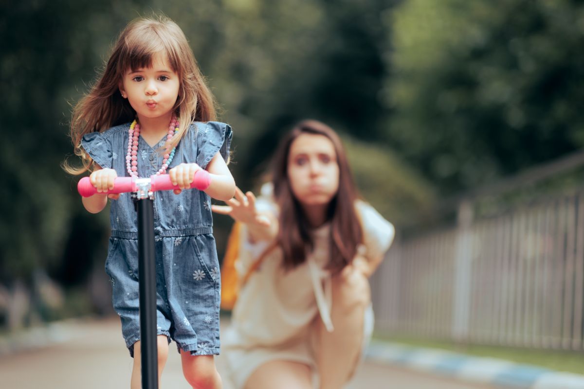 Fetiță, îmbrăcată cu rochiță din jeans, pe trotinetă, în parc, iar mama ei stă în spatele ei și pare foarte îngrijorată, ilustrând 5 temeri ale părinților