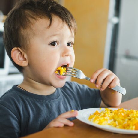 Băiețel care stă la masă, în bucătărie și mănâncă o omletă, cu furculița, dintr-o farfurie albă, iar în spate se poate observa un cuptor cu micounde, ilustrând alimente care provoacă alergii