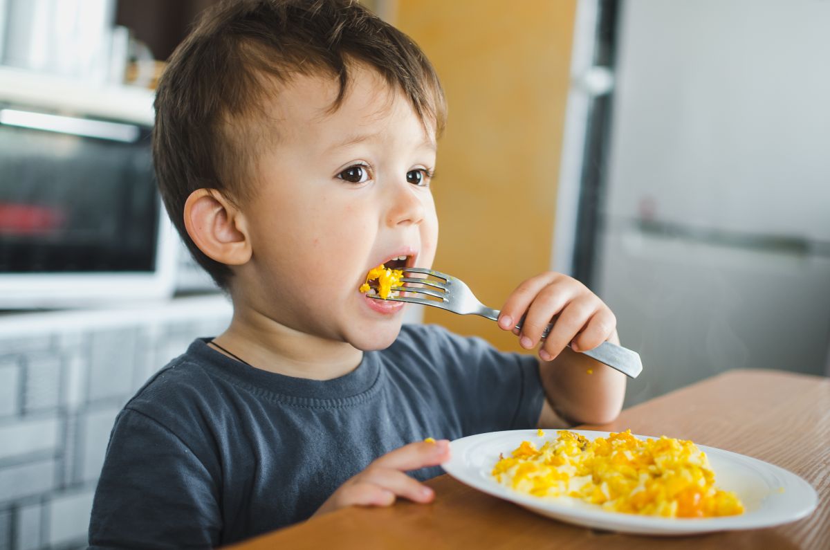 Băiețel care stă la masă, în bucătărie și mănâncă o omletă, cu furculița, dintr-o farfurie albă, iar în spate se poate observa un cuptor cu micounde, ilustrând alimente care provoacă alergii