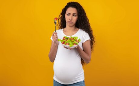 Femeie însărcinată, îmbrăcată cu un tricou alb și jeanși, fotografiată pe un fundal galben, care ține în mână un bol cu salată și o fuculiță rificată, în care este prinsă o roșie, ilustrând aversiunile alimentare în sarcină