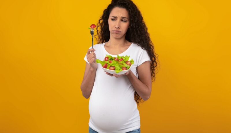 Femeie însărcinată, îmbrăcată cu un tricou alb și jeanși, fotografiată pe un fundal galben, care ține în mână un bol cu salată și o fuculiță rificată, în care este prinsă o roșie, ilustrând aversiunile alimentare în sarcină