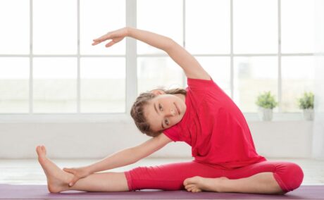 Fetiță, îmbrăcată cu tricou și colanți roșii, într-un studio cu geamuri mari, cu rame albe, practică yoga pe o saltea mov, ținând un picior întins în lateral și celălalt îndoit, cu corpul înclinat spre piciorul întins, cu o mână cuprinzând piciorul întins, iar cealaltă ridicată deasupra capului