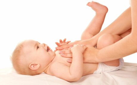 Bebeluș în scutec, care stă pe spate, pe un pat cu cearșaf alb, și îi atinge mamei mâinile, în timp ce aceasta îi face masaj pe piept, cu mâinile