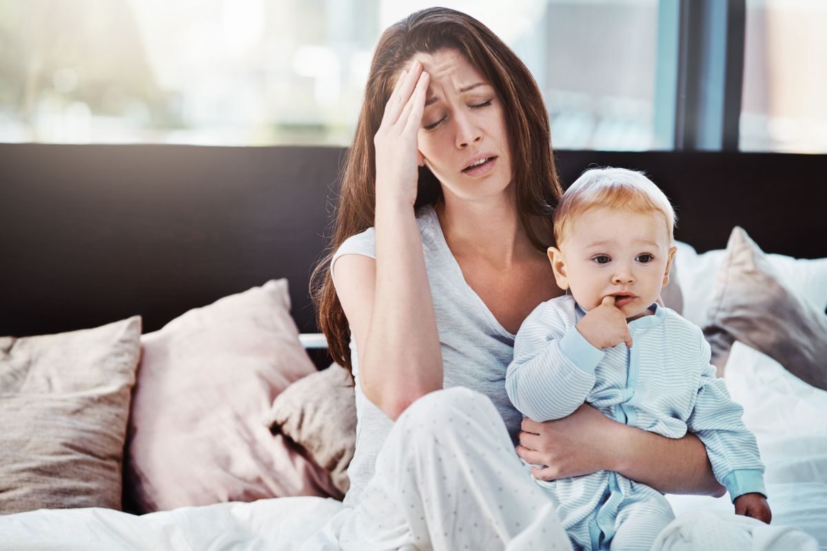 Mamă, care stă pe un pat cu spătar maro, cu multe perne în spate, îmbrăcată cu o pijama bej, își ține o mână la cap și bebelușul în cealaltă mână, ilustrând burnout-ul mamei
