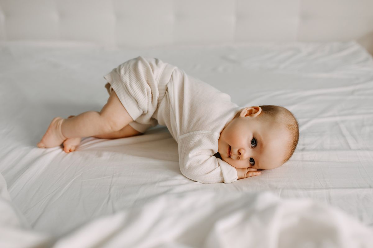 Bebeluș, îmbrăcat cu body alb și pantaloni scurți care stă pe un pat de adulți, acoperit cu cearșaf alb, și încearcă să se rostogolească, stând sprijinit cu capul pe brațe și se sprijină pe un genunchi, având posteriorul ridicat, ilustrând când încep bebelușii să se rostogolească