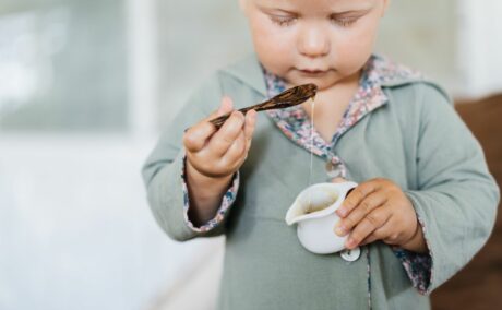 Băiețel, îmbrăcat cu o bluză gri, care ține în mână o cană albă, cu miere și în cealaltă o linguriță din lemn, când introduci mierea în alimentația bebelușului