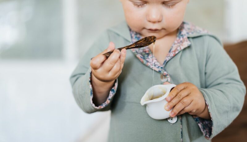 Băiețel, îmbrăcat cu o bluză gri, care ține în mână o cană albă, cu miere și în cealaltă o linguriță din lemn, când introduci mierea în alimentația bebelușului