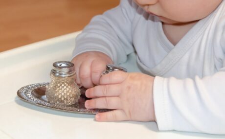 Bebeluș, îmbrăcat în body alb, care stă la masă de bebeluși și ține în mâini o solniță cu sare, iar pe masă mai este o farfurie din metal argintiu și pe ea este așezată o solniță cu piper, ilustrând când poate bebelușul să mănânce sare