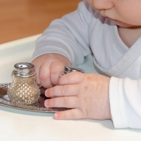 Bebeluș, îmbrăcat în body alb, care stă la masă de bebeluși și ține în mâini o solniță cu sare, iar pe masă mai este o farfurie din metal argintiu și pe ea este așezată o solniță cu piper, ilustrând când poate bebelușul să mănânce sare