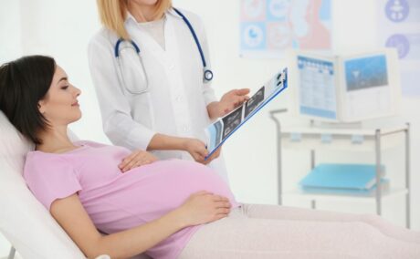 Femeie însărcinată, îmbrăcată cu tricou roz și colanți bej, care stă întinsă pe pat, în cabinetul ginecologului pentru a-i face profilul biofizic, iar medicul în halat alb, cu un stetoscop albastru de gât, îi arată o fișă