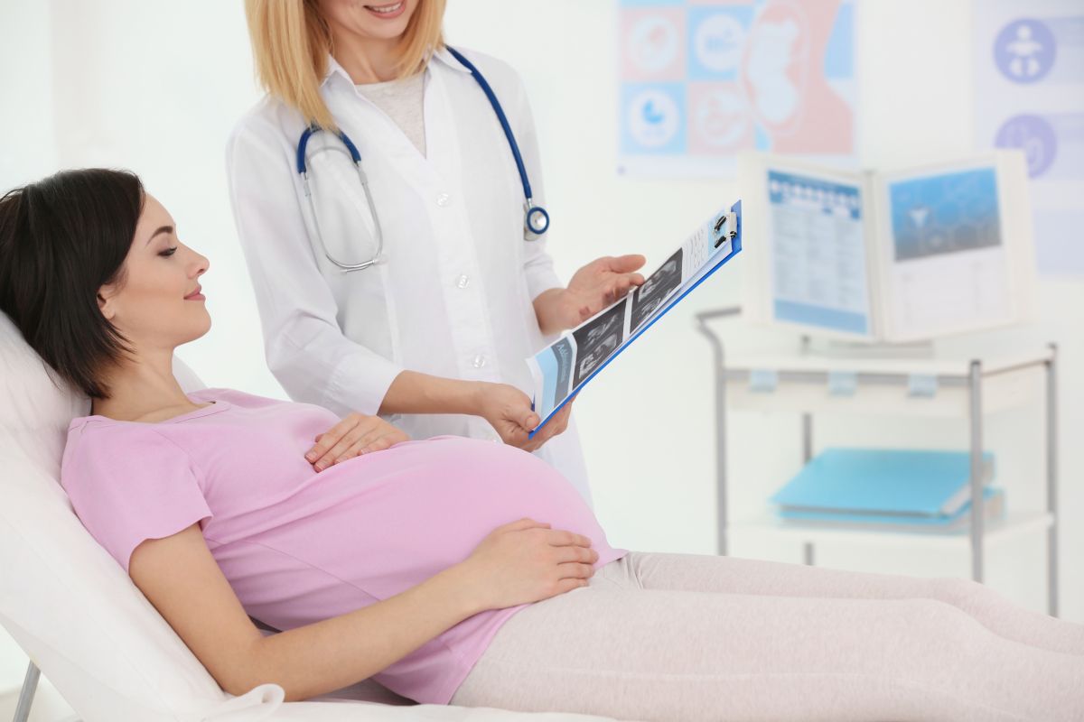 Femeie însărcinată, îmbrăcată cu tricou roz și colanți bej, care stă întinsă pe pat, în cabinetul ginecologului pentru a-i face profilul biofizic, iar medicul în halat alb, cu un stetoscop albastru de gât, îi arată o fișă